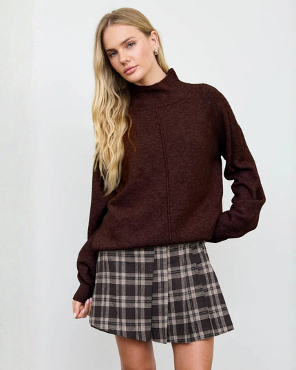 A woman modeling a dark brown mock neck knit sweater paired with a brown and beige plaid pleated mini skirt, set against a plain light gray studio background.