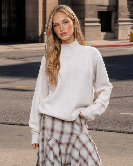 A woman stands on a city street wearing a cream mock-neck knit sweater and a beige-and-white plaid skirt.