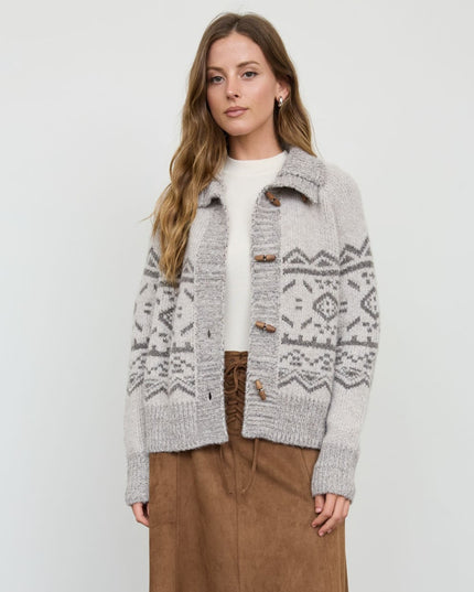 A woman modeling a light gray knit cardigan with a darker gray Fair Isle pattern, brown buttons, and a white top, paired with brown pants, against a plain light gray background.