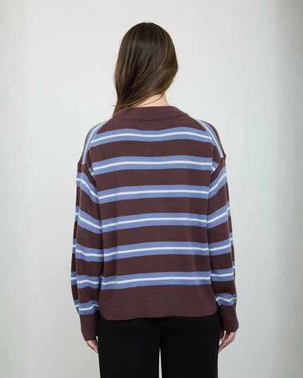A woman viewed from behind wearing a brown knit sweater with horizontal light blue and white stripes, paired with black pants, against a plain light gray background.