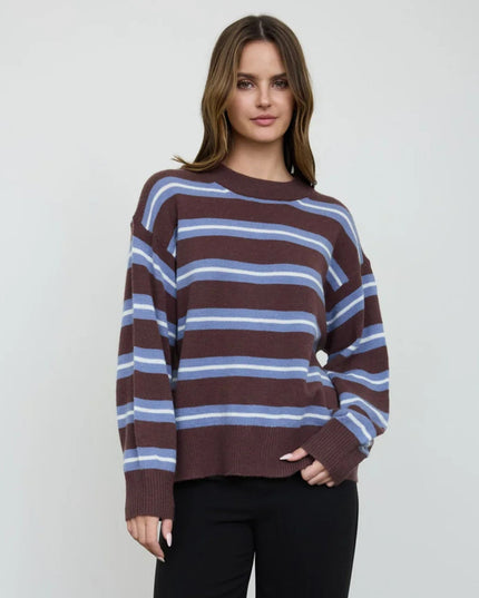 A woman modeling a brown knit sweater with horizontal light blue and white stripes, paired with black pants, against a plain light gray background.