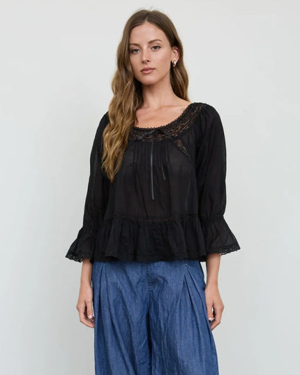 A woman modeling a black sheer long-sleeve blouse with a lace-trimmed scoop neckline and ruffled hem, paired with blue wide-leg pants, against a plain light gray background.