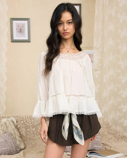 A woman stands indoors wearing a loose white blouse with lace trim and ruffled sleeves, paired with dark brown shorts and a patterned silk tie at the waist. The room has beige floral wallpaper, framed wall art, and a sofa with pillows in the background.