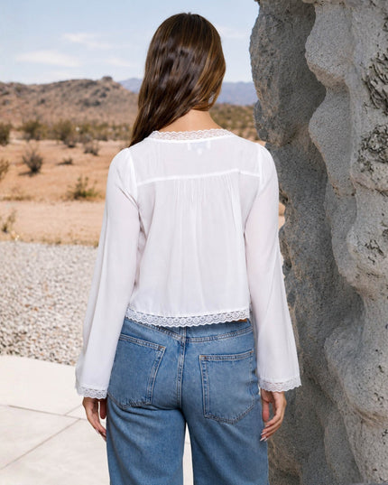 A woman viewed from behind wearing a white long-sleeve sheer blouse with lace trim and blue jeans, standing outdoors next to a large rock formation with a desert landscape in the background.