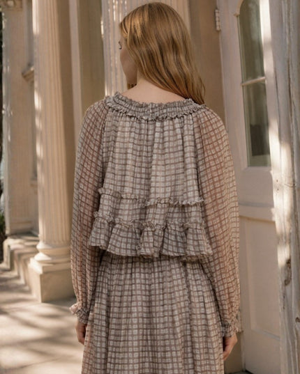 A woman viewed from behind wearing a beige and brown plaid long-sleeve dress with a ruffled back detail, standing in front of a sunlit beige building with columns.