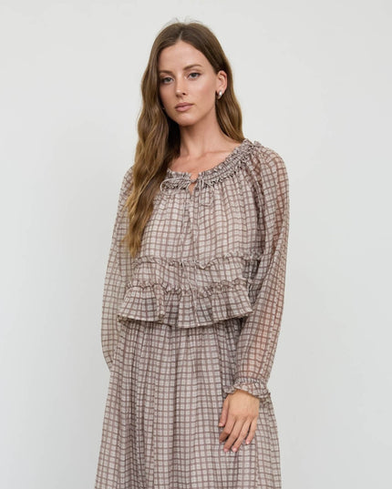 A woman modeling a long-sleeve tiered dress with a brown and beige plaid pattern, set against a plain light gray studio background.