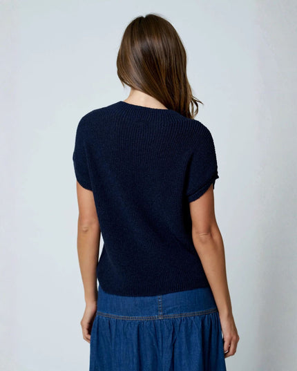 A woman viewed from behind wearing a dark navy ribbed short-sleeve knit top and a blue denim skirt, standing against a plain light gray wall.