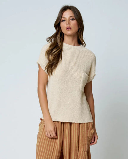 A woman modeling a short-sleeve cream ribbed knit top with a side pocket, paired with brown and beige vertically striped trousers, against a light gray studio background.