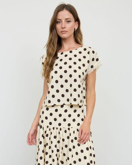 A woman modeling a cream-colored two-piece set with dark brown polka dots, featuring a short-sleeve top with ruffled trim and matching tiered skirt, against a plain light gray background.