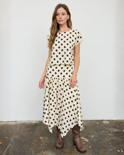 A woman modeling a cream-colored two-piece set with black polka dots, consisting of a short-sleeve top and a matching midi skirt, paired with brown leather boots, standing against a white wall.