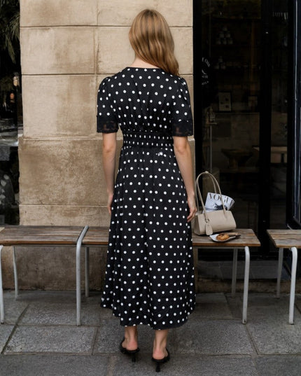 A woman stands with her back to the camera wearing a black midi dress with white polka dots. She is standing on a stone sidewalk in front of a building, next to two wooden benches and a beige handbag.