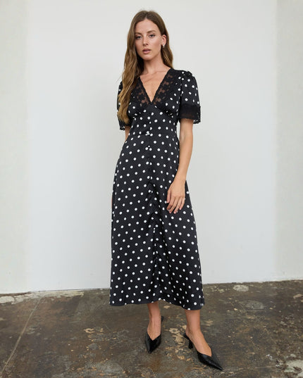 A woman models a black midi dress with white polka dots, featuring short puff sleeves and a V-neckline, standing against a white wall.