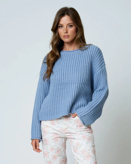 A woman modeling a light blue chunky ribbed knit sweater with balloon sleeves, paired with light floral print pants, against a plain light gray studio background.