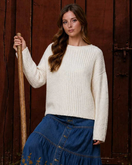 A woman stands in front of a rustic dark wooden door holding a wooden pole, wearing a cream-colored chunky cable-knit sweater and a blue denim skirt.