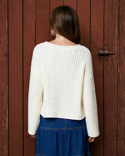 A woman viewed from behind wearing a cream ribbed knit sweater and a blue denim skirt, standing in front of a rustic reddish-brown wooden door.