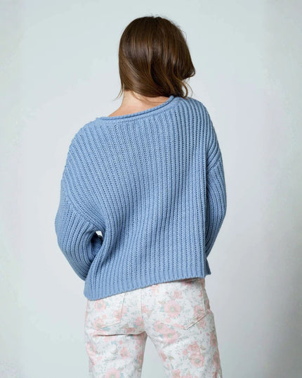 A woman viewed from behind wearing a light blue ribbed knit sweater and light floral pants against a plain light gray background.