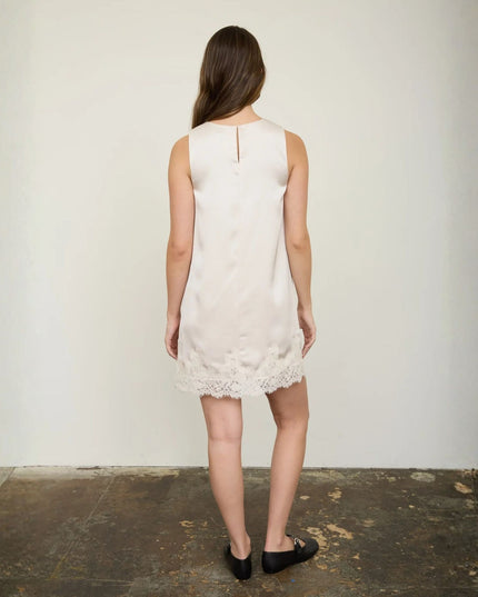 A woman viewed from behind wearing a sleeveless cream-colored dress with a lace hem, standing against a plain off-white wall on a dark floor.
