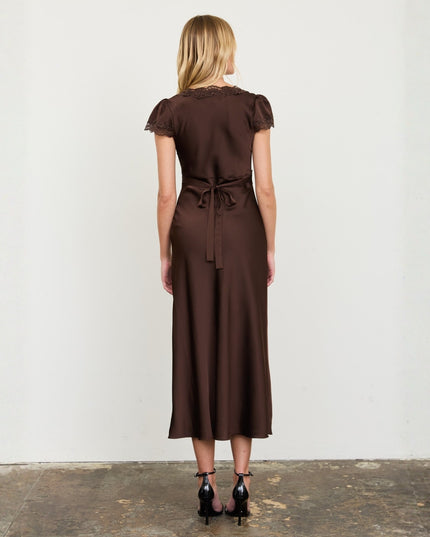 A woman stands with her back to the camera wearing a dark brown midi dress with short flutter sleeves, lace trim at the neckline, and a tie waist, paired with black high heels against a plain off-white wall.