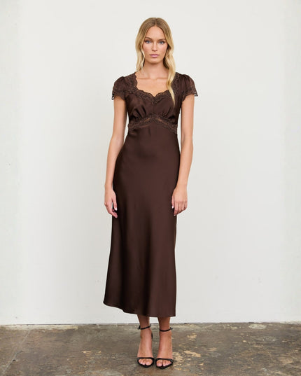 A woman modeling a dark brown midi-length satin slip dress with lace detailing on the bodice and cap sleeves, standing against a plain off-white wall.