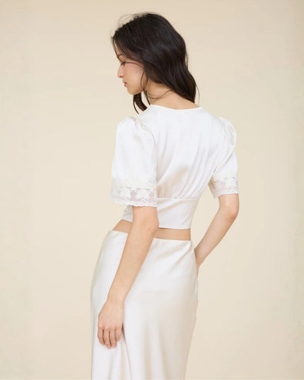A woman shown from the back wearing a white short-sleeve satin crop top with lace trim and a matching white satin skirt against a plain beige studio background.