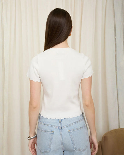 A woman viewed from behind wearing a fitted white short-sleeve top with scalloped edges and light blue jeans, standing in front of cream curtains.