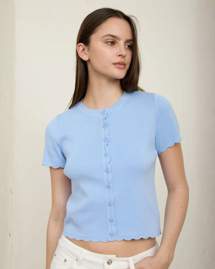 A woman wearing a light blue short-sleeve knit top with scalloped edges and a button front, paired with white pants, standing against a plain off-white wall.