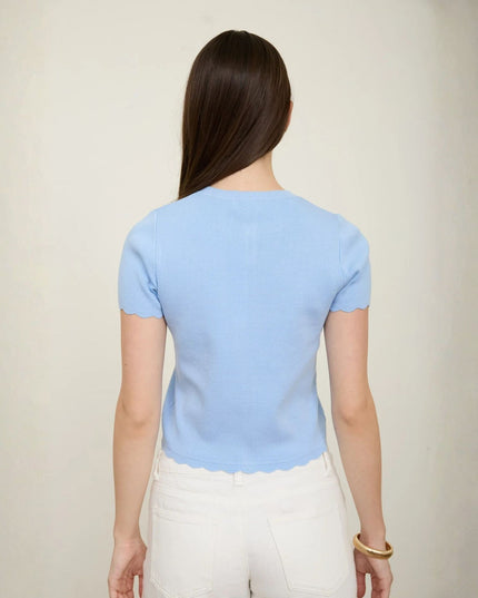 A woman viewed from the back wearing a light blue short-sleeve top with scalloped edges and white pants, standing against a plain light beige wall.
