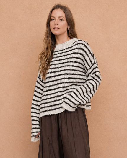 A woman wearing a black and white horizontally striped fuzzy knit sweater and dark brown wide-leg pants, standing against a solid warm beige background.