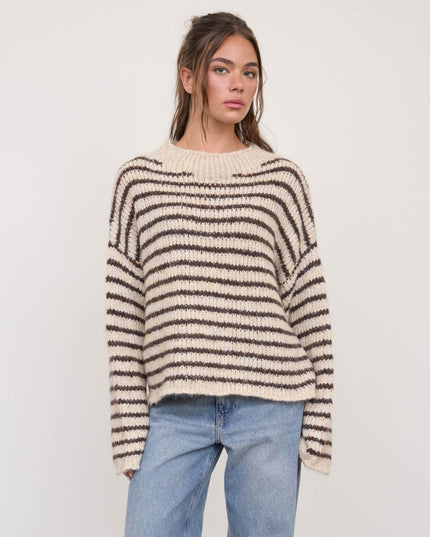 A woman wearing a cream and dark brown horizontally striped knit sweater with light blue jeans, standing against a plain light gray studio background.