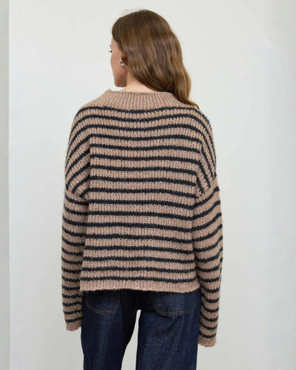 A woman viewed from behind wearing a loose, ribbed knit sweater with horizontal beige and black stripes, paired with dark blue jeans, against a plain off-white wall.