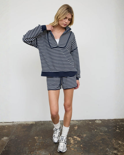 A woman modeling a matching navy and white striped lounge set consisting of a half-zip hoodie and shorts, paired with white socks and silver sneakers, standing against a white wall.
