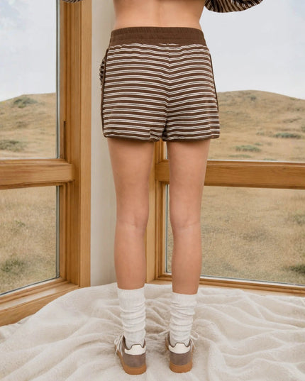 A person standing indoors by large wood-framed windows, wearing brown and beige horizontally striped knit shorts, white socks, and brown shoes, with a dry grassy landscape visible outside.