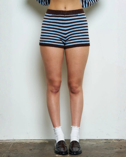 A person from the waist down wearing striped knit shorts with a brown waistband and hem, styled with white socks and dark brown loafers against a plain off-white wall.