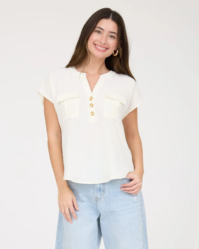 A woman modeling a short-sleeve cream-colored blouse with a split neckline, three gold buttons, and two chest pockets, paired with light blue jeans against a plain white studio background.