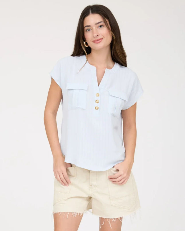 A woman modeling a light blue short-sleeve button-front top with two chest pockets, paired with light beige shorts, against a plain white studio background.