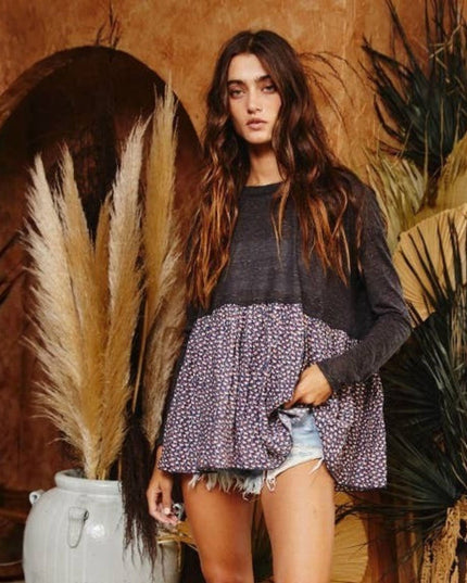 A woman modeling a long-sleeve color-block top with a dark charcoal upper section and a purple-and-white patterned flared lower section, styled with denim shorts in a rustic boho setting with dried grass and palm leaves.