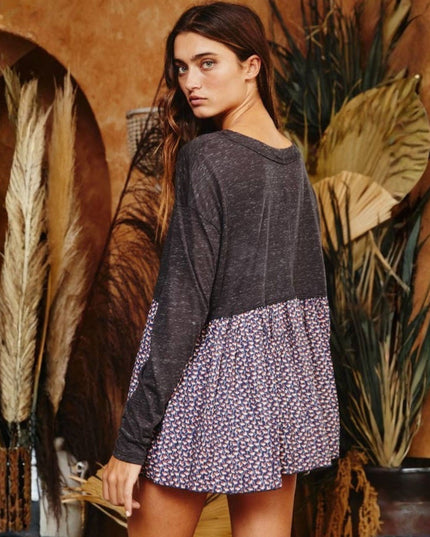 A woman wearing a long-sleeve colorblock top with a dark charcoal gray upper section and a purple, pink, and white floral patterned skirt, posed in front of a rustic brown wall with dried plants.