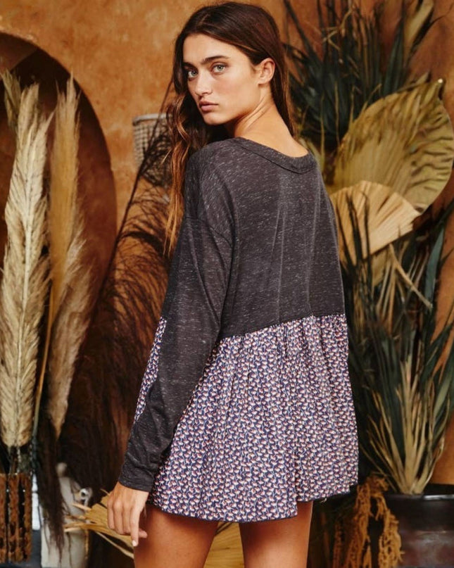 A woman wearing a long-sleeve colorblock top with a dark charcoal gray upper section and a purple, pink, and white floral patterned skirt, posed in front of a rustic brown wall with dried plants.