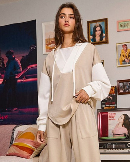 A woman models a beige and white color-blocked hoodie and matching sweatpants set in a styled room with framed pictures and a record player in the background.