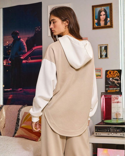 A woman wearing a beige and white color-blocked hoodie and matching sweatpants, photographed from behind in a cozy, decorated room with framed pictures and a record player.