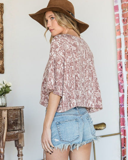 A woman wearing a dusty rose and white patterned short-sleeve blouse with denim shorts and a brown wide-brim hat, standing in a decorated room with a wooden table and mirror.