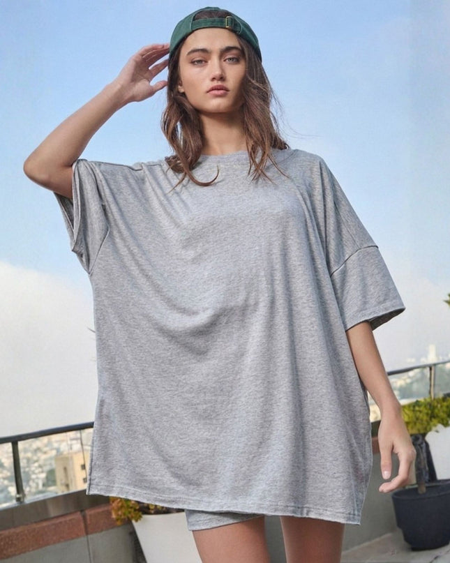 A woman wearing an oversized light gray heather t-shirt and matching shorts, with a green baseball cap backwards, standing on a rooftop terrace with a city skyline in the background.