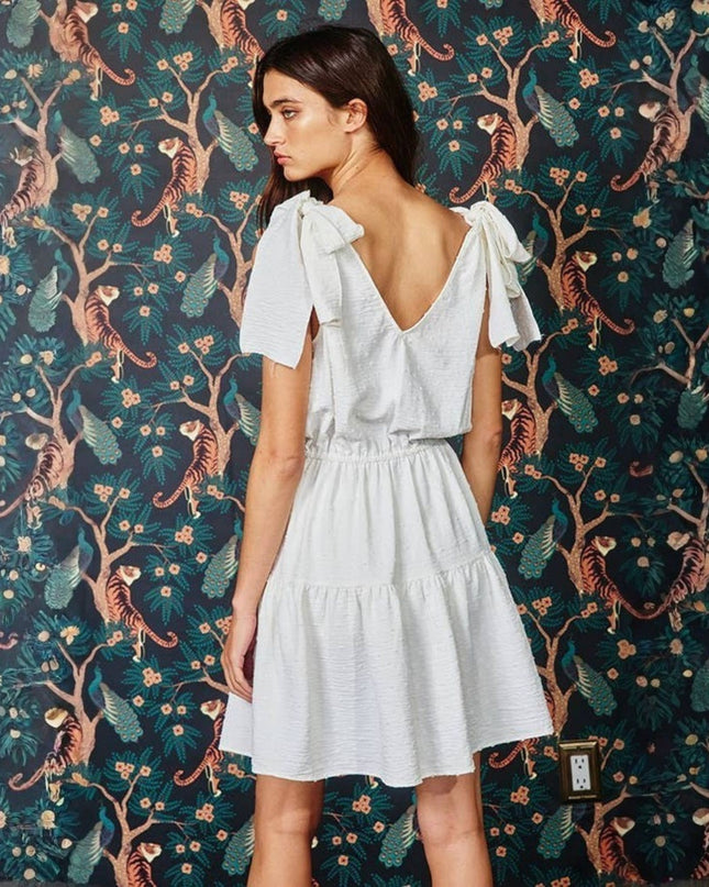 A woman wearing a white tiered mini dress with ruffled short sleeves and a deep V-back, standing in front of a dark green and brown jungle-patterned wallpaper.
