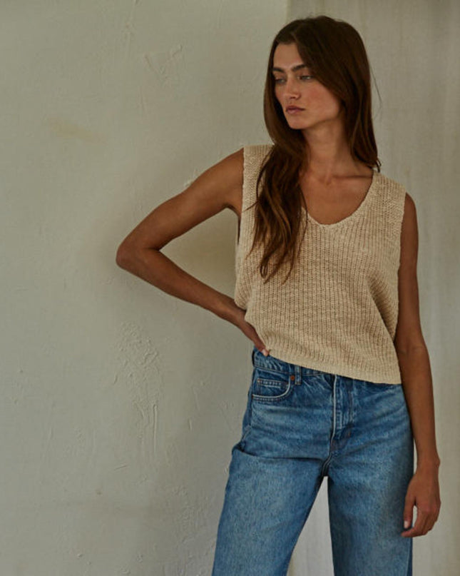Woman wearing a beige sleeveless top and blue jeans against a plain wall.