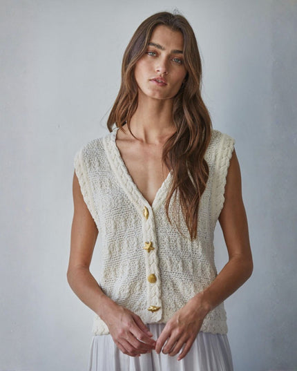 Woman wearing a cream-colored knitted vest with gold buttons against a plain background