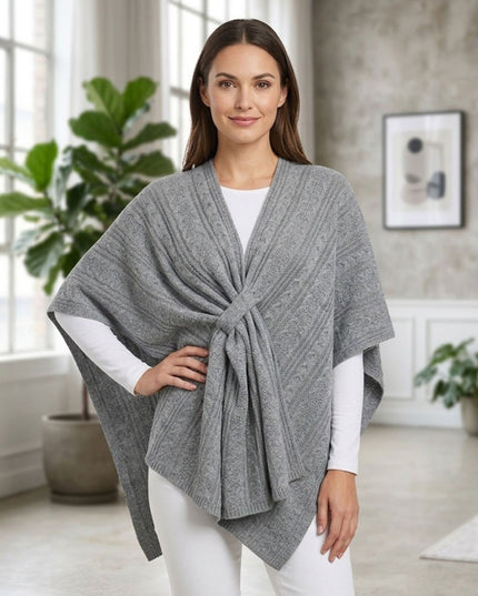 A woman modeling a gray cable-knit wrap poncho over a white long-sleeve top and white pants, standing in a bright, modern room with potted plants and a textured wall.