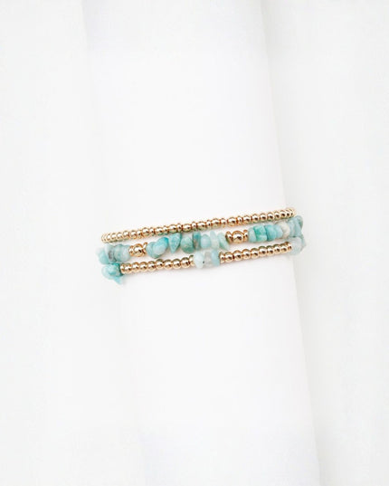 A pair of double-strand beaded bracelets featuring light blue-green chunky stones and small gold metallic beads, displayed on a white cylindrical stand.