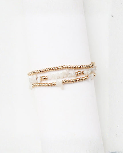 Three delicate beaded bracelets stacked on a white cylindrical display stand. The bracelets feature metallic rose gold beads and translucent white stone beads.