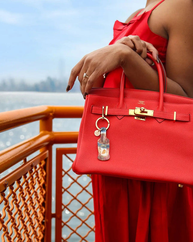 A woman in a red dress holds a bright red structured handbag with gold-tone hardware and a hanging keychain, standing by an orange railing over water.