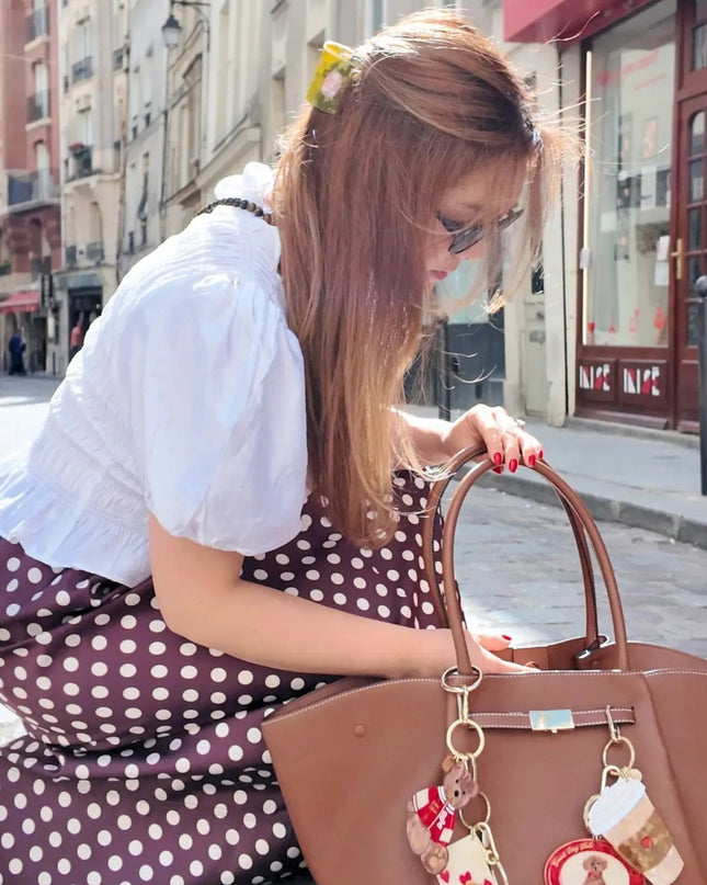 A woman in a white puff-sleeve blouse and a brown polka-dot skirt is crouching on a cobblestone street, holding a large brown leather handbag with gold-tone hardware and hanging charms.